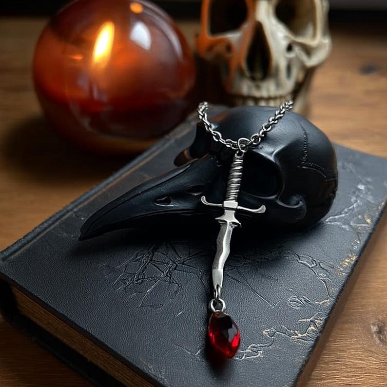 May include: A silver dagger pendant with a red gemstone drop hangs from a chain. The pendant rests on a black raven skull atop a dark book. A lit candle and a skull are in the background, creating a gothic aesthetic.
