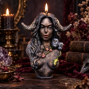 May include: A dark-toned, sculpted candle bust of a figure with long hair, prominent horns, and a neutral expression. The figure is adorned with small pink flowers, two grey owl figurines, and a small green lizard, with a single lit wick at the top.