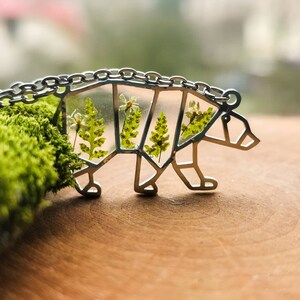 May include: A silver bear necklace with a clear resin inlay featuring pressed green ferns and white flowers.