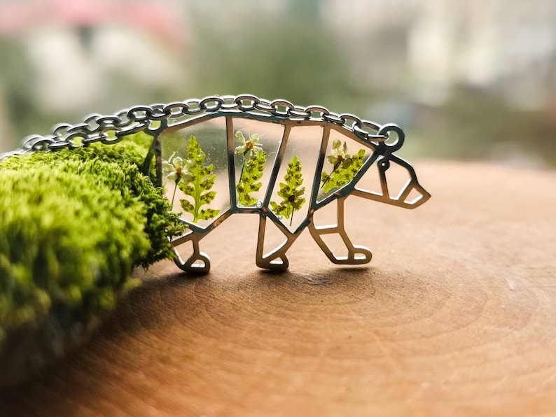 May include: A silver bear necklace with a clear resin inlay featuring pressed green ferns and white flowers.