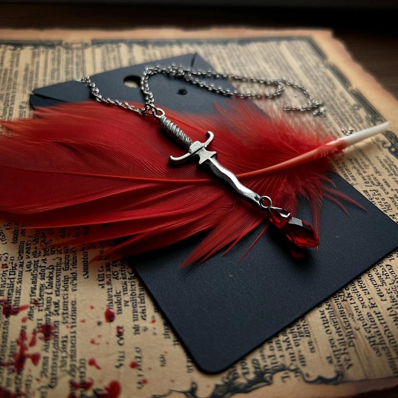 May include: A silver dagger pendant necklace with a red gemstone charm hangs over a vibrant red feather. The necklace rests on a black card, set against a backdrop of aged paper with text and a dark surface.