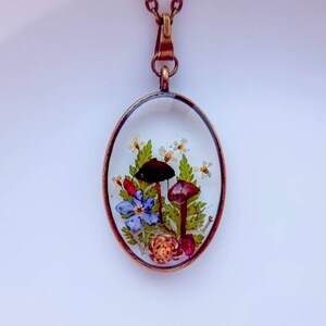 May include: A delicate oval pendant necklace with a clear resin setting featuring pressed flowers, mushrooms, and a small snail. The pendant is attached to a bronze chain.