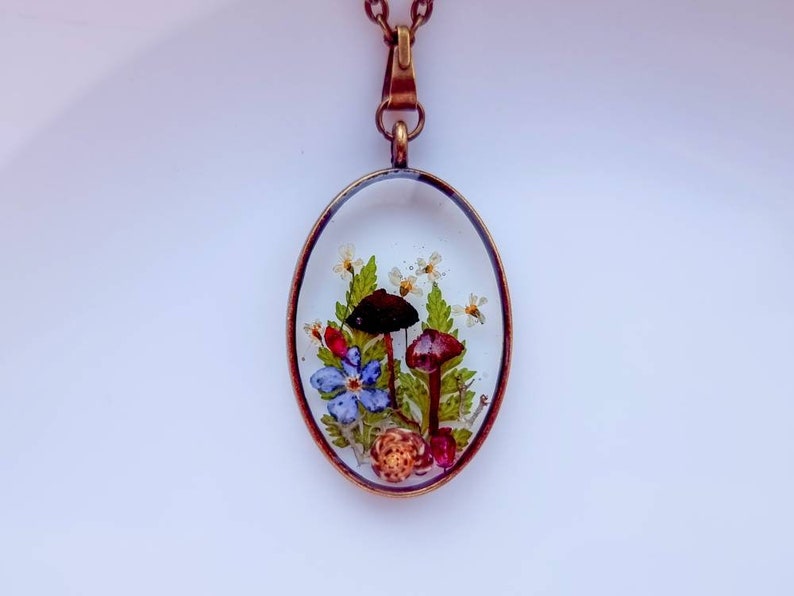 May include: A delicate oval pendant necklace with a clear resin setting featuring pressed flowers, mushrooms, and a small snail. The pendant is attached to a bronze chain.