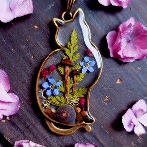 May include: A gold cat-shaped pendant necklace with a clear resin insert containing dried flowers, ferns, mushrooms, and a snail shell.