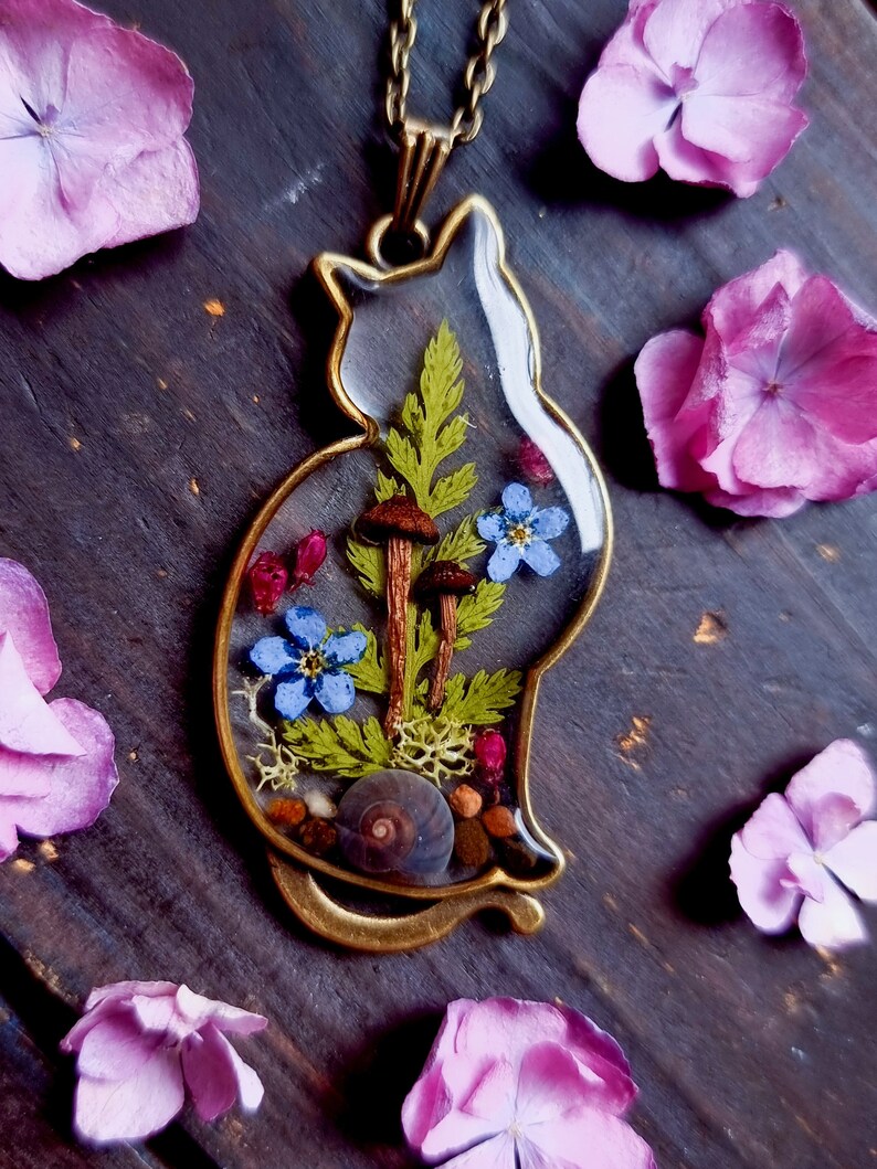 May include: A gold cat-shaped pendant necklace with a clear resin insert containing dried flowers, ferns, mushrooms, and a snail shell.