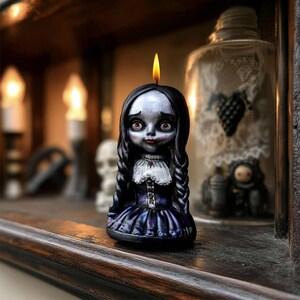 May include: A doll-shaped candle with a lit wick. The candle features a pale face, dark hair, and a dark blue dress with a white collar. The candle is on a wooden shelf with other decorative items.