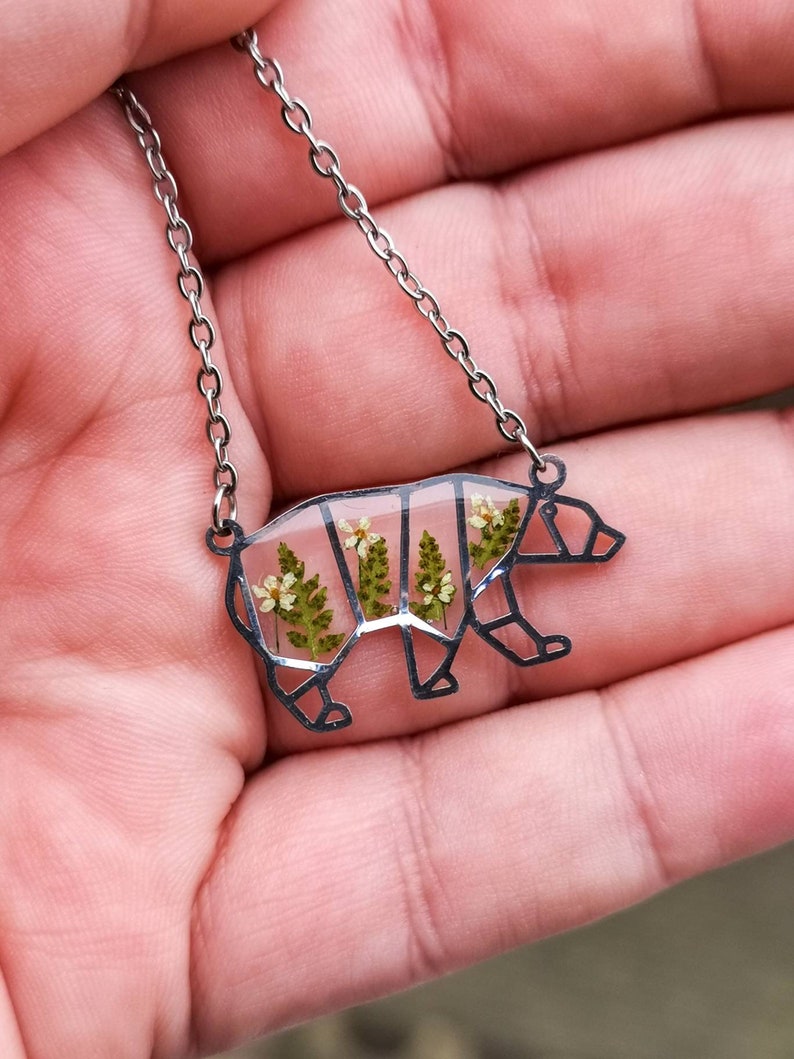 May include: A silver chain necklace with a clear resin pendant shaped like a bear. The pendant contains pressed flowers and ferns.