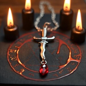 May include: A silver pendant necklace featuring a dagger design with a red gemstone. The pendant is displayed on a dark surface with a red circular design and surrounded by four lit black candles.