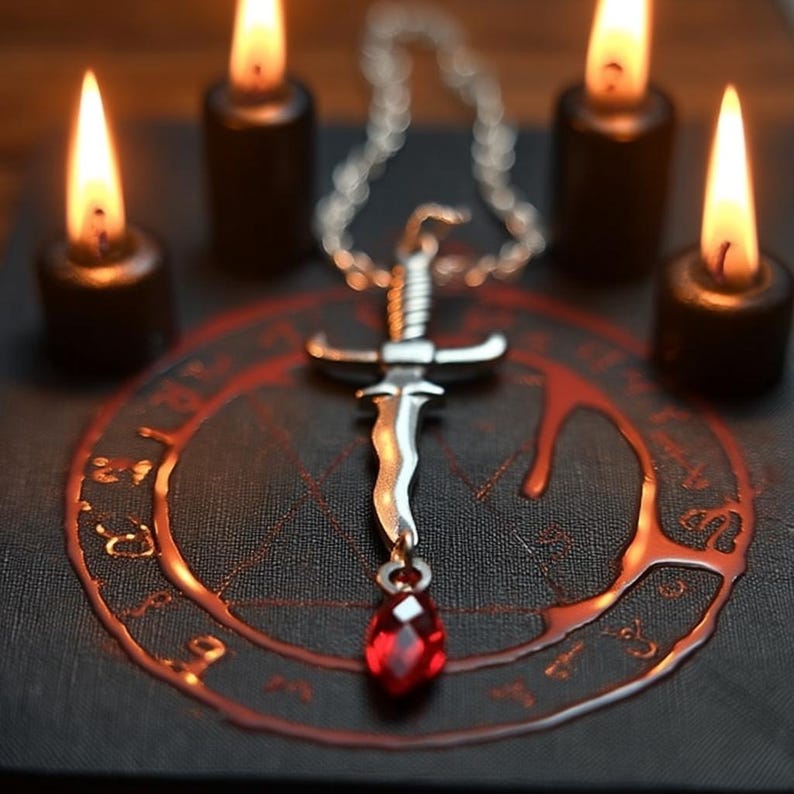 May include: A silver pendant necklace featuring a dagger design with a red gemstone. The pendant is displayed on a dark surface with a red circular design and surrounded by four lit black candles.