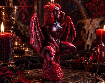 May include: A dark red wax candle shaped as a winged figure with horns and a serpent coiled around its base. The figure has long hair, a pointed tail, and a small symbol on its chest. A single flame burns from the top of the figure's head.