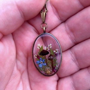 May include: A delicate oval pendant necklace with a clear resin charm featuring pressed flowers and mushrooms. The charm is set in a bronze frame and hangs from a bronze chain.