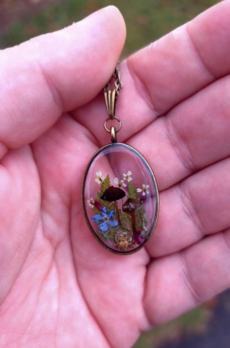 May include: A delicate oval pendant necklace with a clear resin charm featuring pressed flowers and mushrooms. The charm is set in a bronze frame and hangs from a bronze chain.