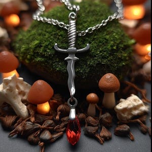 May include: A silver-colored pendant necklace featuring a miniature sword design with a red gemstone at the bottom. The sword is suspended from a chain and set against a backdrop of moss, mushrooms, and other natural elements.