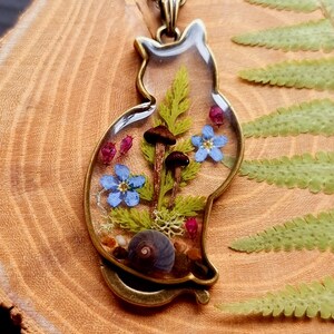 May include: A clear resin cat pendant necklace with a bronze chain. The pendant contains pressed flowers, mushrooms, and a snail.