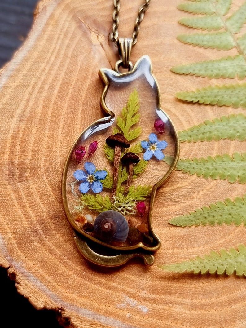 May include: A clear resin cat pendant necklace with a bronze chain. The pendant contains pressed flowers, mushrooms, and a snail.