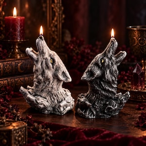 May include: Two sculpted wax candles shaped like wolf heads howling at the moon. One candle is white with grey shading, and the other is black with silver highlights. Both feature yellow eyes and small wolf figures at the base, with a lit wick on top.