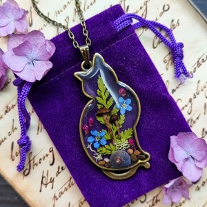May include: A bronze pendant necklace featuring a cat silhouette filled with pressed flowers, mushrooms, and greenery. The pendant is on a purple velvet pouch.