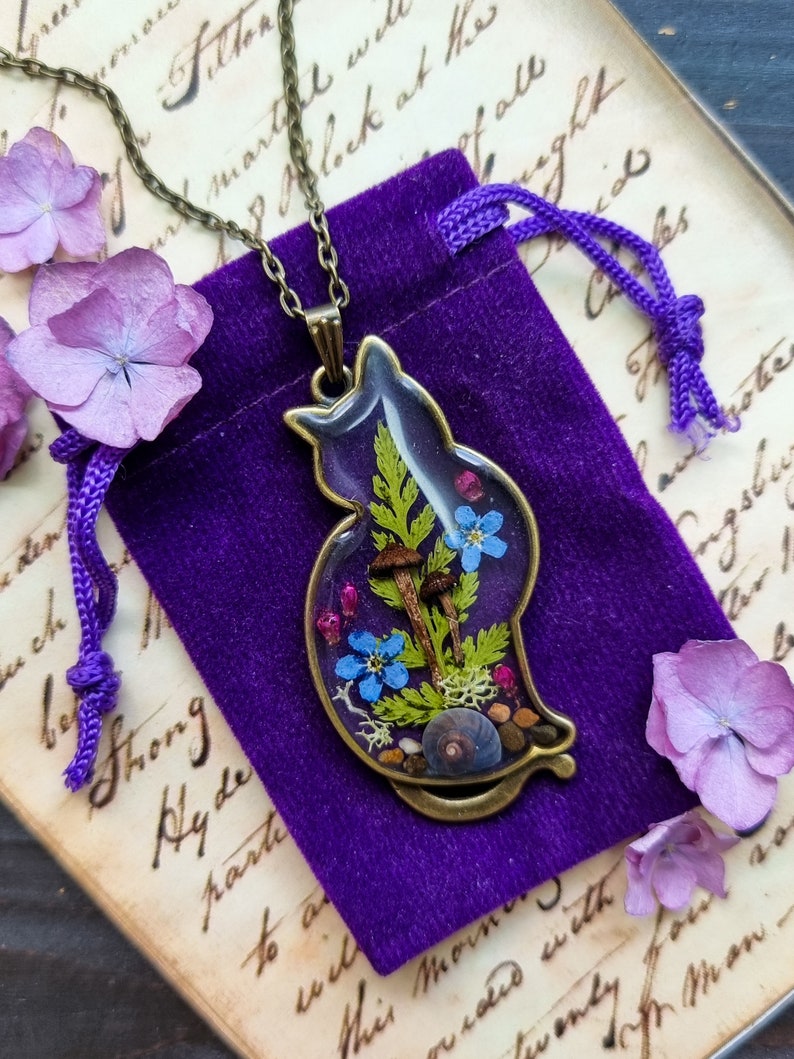 May include: A bronze pendant necklace featuring a cat silhouette filled with pressed flowers, mushrooms, and greenery. The pendant is on a purple velvet pouch.