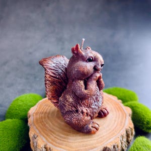 Puede incluir: Una vela de ardilla marr&oacute;n con un dise&ntilde;o detallado, sentada sobre una rodaja de madera. La ardilla est&aacute; sosteniendo sus patas hacia su cara, como si estuviera pensando.