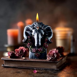 May include: A black ram-shaped candle with a lit wick, adorned with a red gem and floral accents. The candle sits atop a vintage book, surrounded by dried roses, creating a gothic and mystical aesthetic.
