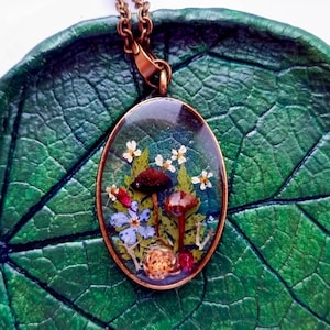 May include: A delicate necklace with a clear oval pendant featuring pressed mushrooms, flowers, and leaves. The pendant is set in a bronze-colored metal frame and hangs from a bronze chain.