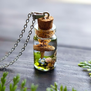 Könnte beinhalten: Eine silberne Kettenkette mit einem kleinen Glasflaschenanhänger. Die Flasche enthält einen Miniaturpilz, Moos und Blumen. Die Flasche hat einen Korkverschluss.