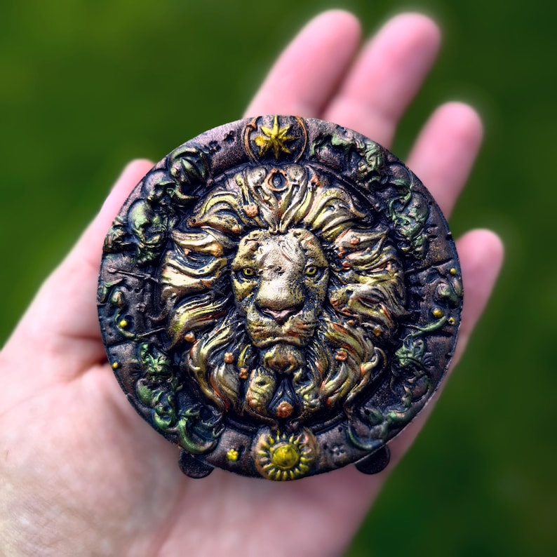 May include: A detailed, round, decorative medallion featuring a golden lion's head at the center. The lion is surrounded by ornate, textured details in shades of gold, brown, and green. The medallion is held in a hand.