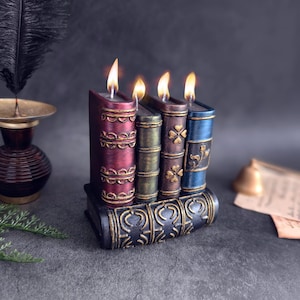 Puede incluir: Cuatro velas decorativas con forma de libros con detalles dorados y llamas encendidas. Las velas están dispuestas en fila sobre una base negra con detalles dorados.