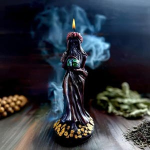May include: A dark purple candle in the shape of a robed figure, holding a green orb. The candle has a red floral crown and a gold base. A lit flame sits atop the figure's head. Smoke and other items are in the background.