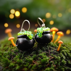 May include: A pair of black cauldron earrings with green bubbling potion detail. The earrings have silver-colored hooks and are set against a backdrop of green moss and miniature orange mushrooms. The background has soft, blurred bokeh lights.
