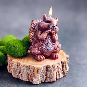 Puede incluir: Una vela de ardilla marr&oacute;n y blanca con una mecha encendida, sentada sobre una rodaja de tronco de madera. La vela est&aacute; detallada con pelaje y tiene un aspecto realista.