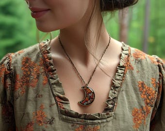May include: A bronze-toned necklace with a crescent moon pendant. The pendant is filled with a colorful floral design. The woman is wearing a green dress with orange floral patterns. The background is a blurred green.