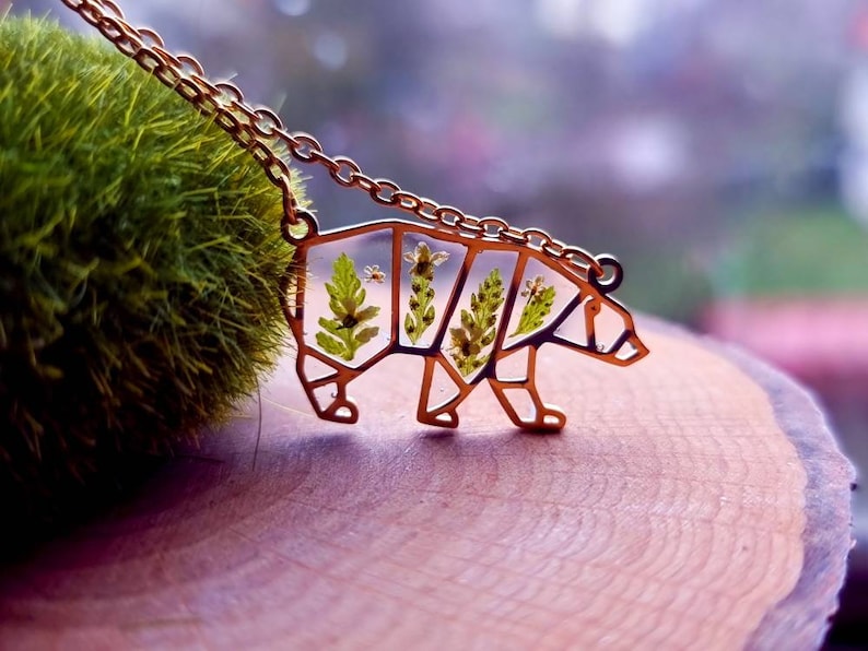 May include: A gold-toned necklace featuring a bear pendant. The bear is crafted from metal and has a geometric design. The bear is filled with pressed flowers and leaves.