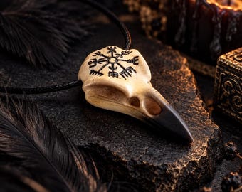 May include: A pendant shaped like a bird skull with a black Vegvisir symbol etched onto the top surface. The skull is cream-colored with a dark, pointed beak and is attached to a thin black cord necklace for wearing around the neck.