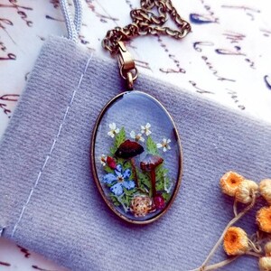 May include: A delicate oval pendant necklace featuring a clear resin design with pressed flowers and mushrooms. The pendant is set in a brass frame and hangs from a gold chain.