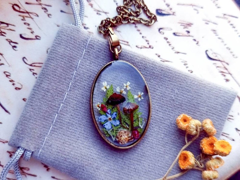 May include: A delicate oval pendant necklace featuring a clear resin design with pressed flowers and mushrooms. The pendant is set in a brass frame and hangs from a gold chain.