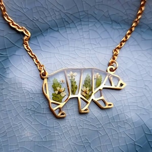 May include: A gold chain necklace with a bear-shaped pendant. The pendant is made of clear resin with pressed flowers inside.