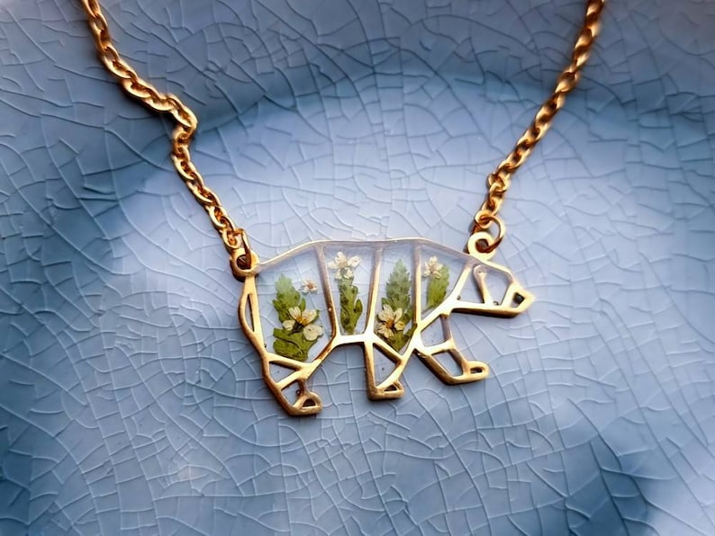 May include: A gold chain necklace with a bear-shaped pendant. The pendant is made of clear resin with pressed flowers inside.