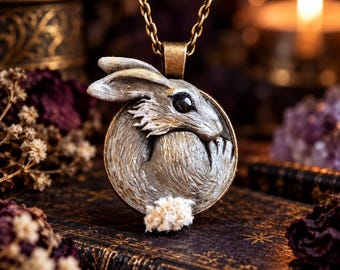 May include: A circular pendant necklace featuring a sculpted rabbit in profile with textured silver and gold-toned fur, a dark eye, and a small white tuft of material at the base, suspended from a metallic chain with a bronze-toned finish.