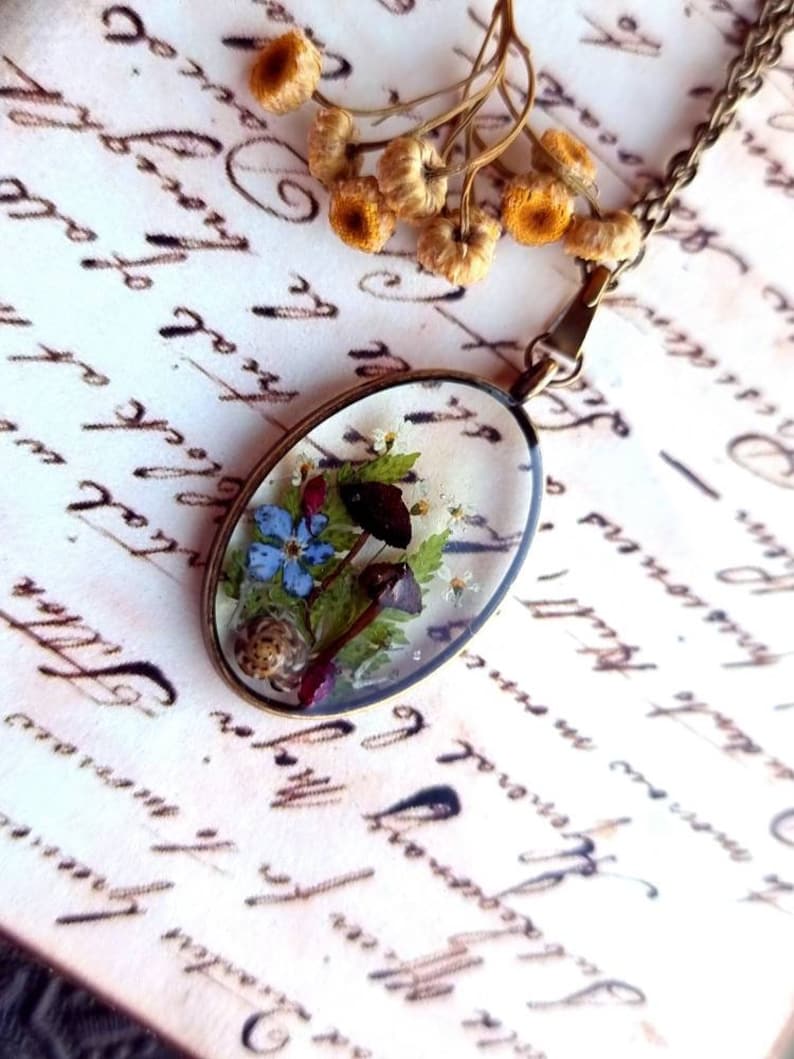May include: A gold chain necklace with a clear oval pendant containing pressed flowers and a small snail. The pendant is on a white background with black writing.