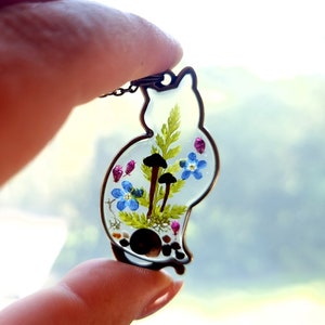 May include: A clear resin pendant shaped like a cat, with a delicate fern, blue forget-me-not flowers, and small brown mushrooms pressed inside. The pendant is on a silver chain.