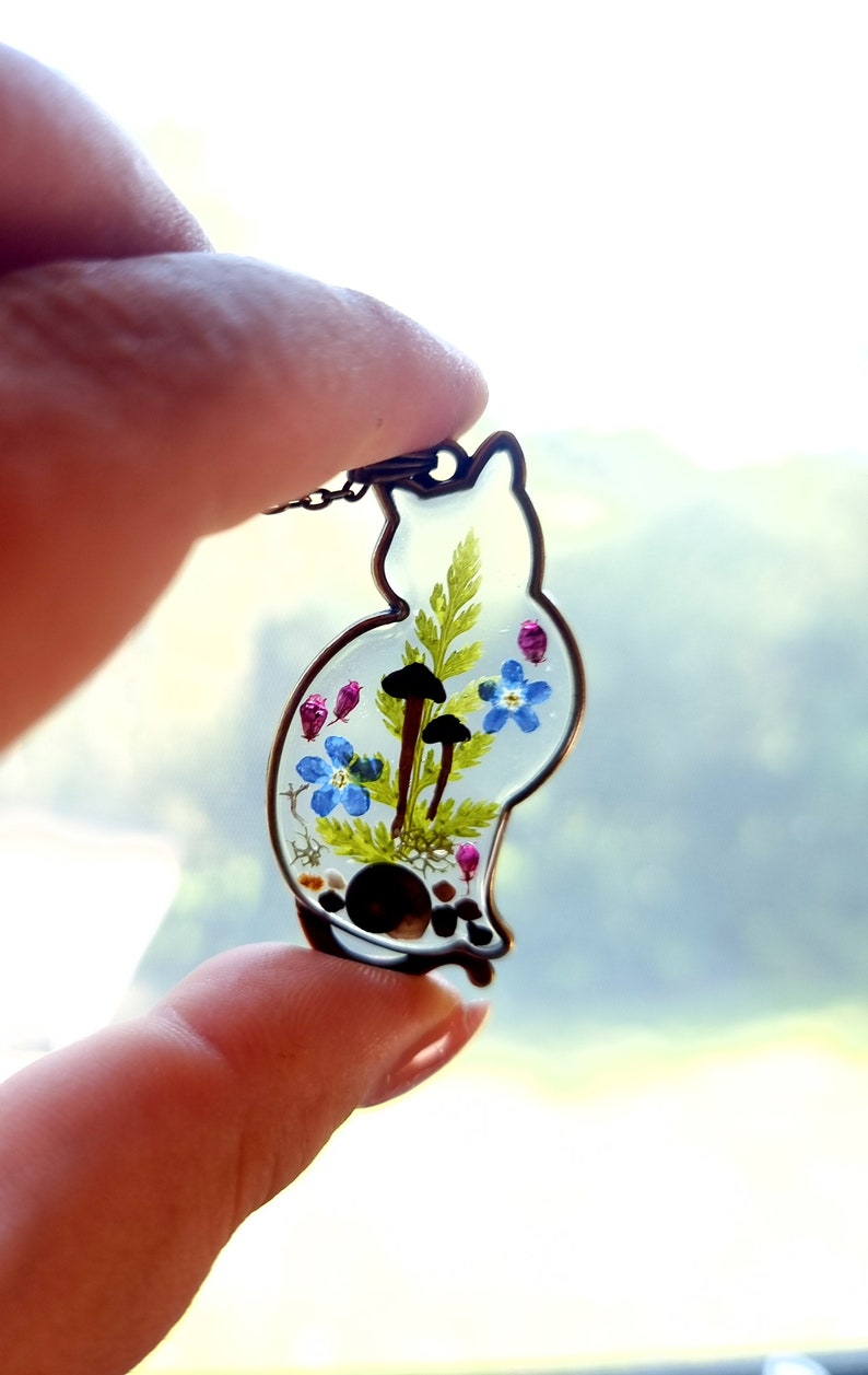 May include: A clear resin pendant shaped like a cat, with a delicate fern, blue forget-me-not flowers, and small brown mushrooms pressed inside. The pendant is on a silver chain.