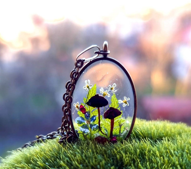 May include: A delicate necklace with a clear glass pendant containing tiny white flowers, green leaves, and two small mushrooms, one black and one brown.