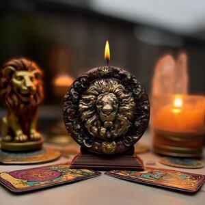 May include: A lit candle with a lion's head design. The candle is brown and gold, with a detailed relief of a lion's face. The candle sits on a wooden base, with tarot cards in the foreground. A golden lion statue is in the background.