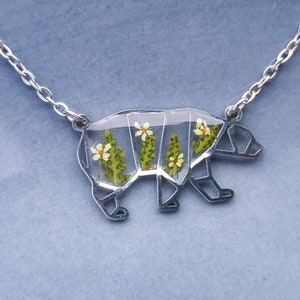 May include: A silver necklace with a clear pendant shaped like a bear. The pendant is filled with pressed flowers and greenery.