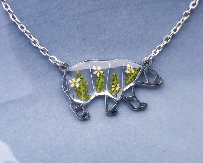 May include: A silver necklace with a clear pendant shaped like a bear. The pendant is filled with pressed flowers and greenery.
