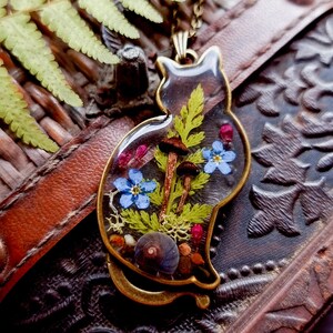 May include: A gold cat-shaped pendant with a clear resin insert containing pressed flowers, ferns, mushrooms, and seashells.