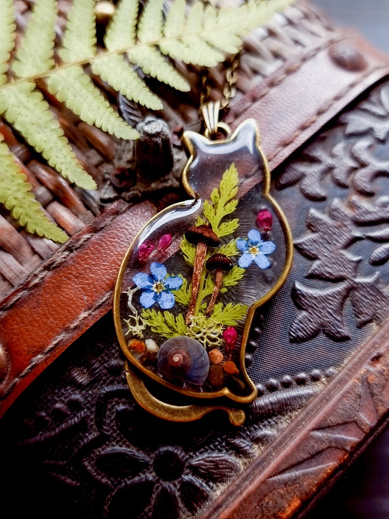 May include: A gold cat-shaped pendant with a clear resin insert containing pressed flowers, ferns, mushrooms, and seashells.