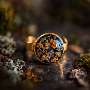 May include: A gold-toned ring with a round, dark blue face. The face is filled with a colorful pattern of orange, yellow, and green organic shapes. The ring band is a simple, adjustable design.