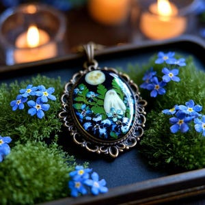 Può includere: Una collana con pendente ovale con una cornice in filigrana color bronzo. Il pendente presenta un disegno di fantasma con luna piena, fiori blu e fogliame verde su sfondo scuro. Fiori blu non ti scordar di me e muschio verde circondano il pendente.